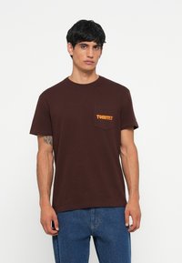 Tricou din bumbac maro cu guler rotund. Are un buzunar pe piept cu logo-ul portocaliu "Tommy". Mâneci scurte și o croială lejeră.
