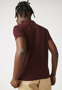 Burgundy polo-skjorta med korta ärmar, ribbat krage och åtsittande design; bärs tillsammans med ljusa byxor. Texturerad tyg synlig.