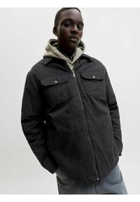 Personne portant une veste noire zippée par-dessus un sweat à capuche beige, les mains dans les poches, sur un fond clair uni.