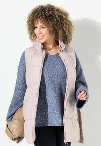 Helles, rosa gepolstertes Weste mit hohem Kragen, über einem blau gesprenkelten Pullover getragen. Hat einen lockeren Schnitt und weite Ärmel, kombiniert mit einer beigen Tasche.