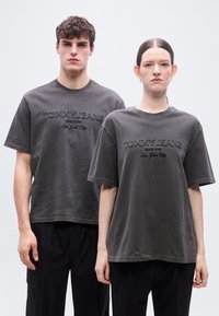 Δύο μοντέλα στέκονται δίπλα-δίπλα φορώντας γκρι μπλουζάκια "Tommy Jeans" και μαύρο παντελόνι, μπροστά σε απλό λευκό φόντο.