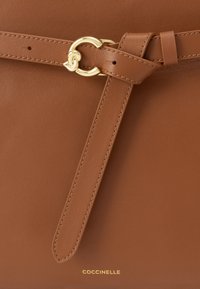 Καφέ δερμάτινη τσάντα χειρός με λεία υφή και χρυσή κυκλική μεταλλική λεπτομέρεια. Διαθέτει διακοσμητικό λουρί και ανάγλυφο λογότυπο στη βάση.