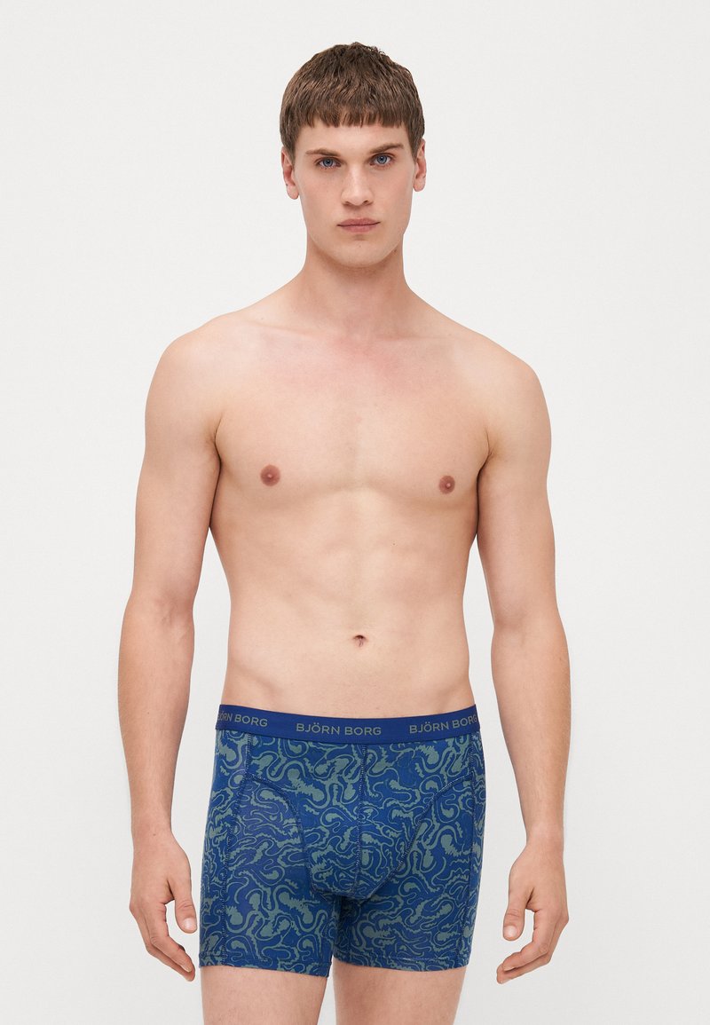Blaue gemusterte Boxer-Shorts mit einem grünen Wirbel-Design, ausgestattet mit einem elastischen, markierten Bund. Das Material wirkt weich und dehnbar.