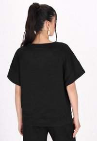 Sort tekstureret bluse med geometrisk mønster, vide ærmer og afslappet pasform. Materialet ser blødt og letvægtigt ud.