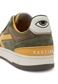 Zapatilla de gamuza verde y amarilla con un talón de cuero blanco que presenta un diseño de ojo, suela de goma texturizada en marrón y detalle en relieve "KAOTIKD".