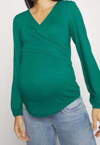 Grön långärmad graviditetsblus med V-ringning, rynkad tyg vid midjan och en slät, mjuk textur. Bärs med ljusblå jeans.