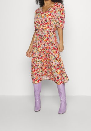 Robe midi à fleurs avec de courtes manches bouffantes, décolleté en V et taille nouée. Présente des couleurs vives et un tissu léger. Associée à des bottes violettes.