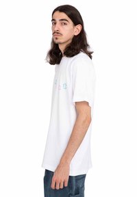 Witte katoenen t-shirt met korte mouwen, voorzien van een meerkleurig tekstontwerp op de voorkant. Model wordt zijwaarts getoond, gekleed in denim broek.