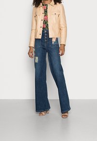 Beige läderjacka, svart blommig topp, och vida blå denimjeans med blommönster. Modell bär bruna klacksandaler.