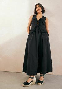 Abito nero senza maniche con dettaglio legato sul davanti, gonna fluida e tasche. Abbinato a espadrillas nere con zeppa e cinturini alla caviglia.