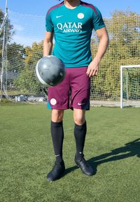 Nike urheilupaita turkoosina, jossa on violetit yksityiskohdat, viininpunaiset shortsit, mustat polvipituiset sukat ja mustat nappulakengät, pitäen harmaata jalkapalloa nurmella.