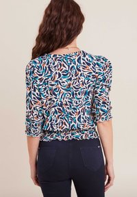 Blusa con motivo floreale, dotata di maniche a palloncino a tre quarti, orlo arricciato e una combinazione di colori blu, arancio e crema su un tessuto leggero.