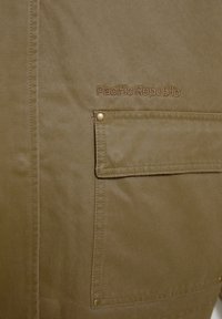 Veste kaki avec un logo brodé "Pacific Republic", une poche avant à fermeture pression et un tissu texturé.