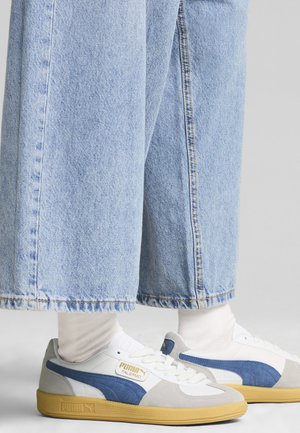 Zapatillas blancas y azules con detalles en ante gris, suela de goma texturizada y diseño de cordones. Combinadas con unos pantalones vaqueros de pierna ancha de color azul claro.