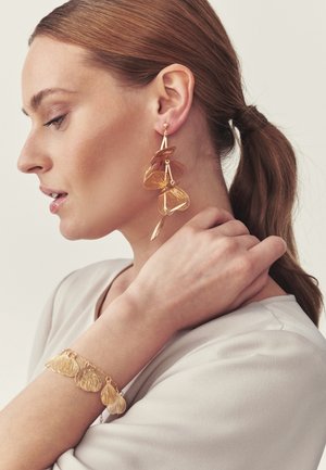 Vrouw met lichtbruin haar in een paardenstaart draagt gouden oorbellen in bladvorm en een bijpassende armband, terwijl ze zachtjes haar hals aanraakt.