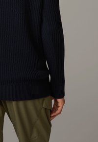 Pull en maille bleu marine avec texture côtelée, manches longues et ourlet à hauteur de la taille. Associé à un pantalon vert olive.