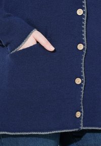Hand in de zak van een donkerblauwe gebreide cardigan met houten knopen en grijze afwerking, gedragen over een spijkerbroek.