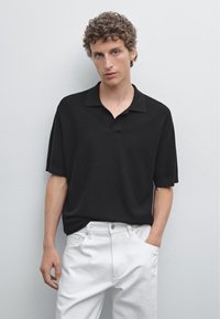 Polo nero con vestibilità rilassata, dotato di colletto e maniche corte, abbinato a pantaloni bianchi. Dettagli in tessuto testurizzato visibili.