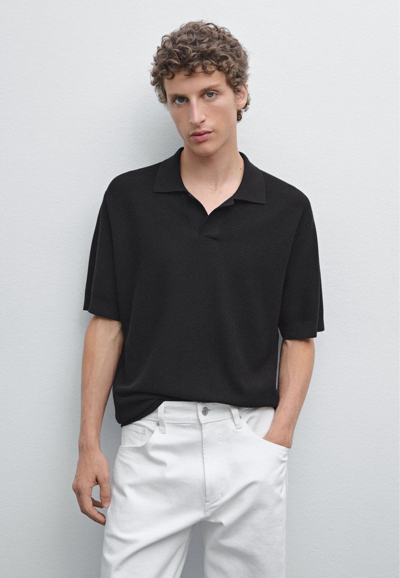 Polo nero con vestibilità rilassata, dotato di colletto e maniche corte, abbinato a pantaloni bianchi. Dettagli in tessuto testurizzato visibili.