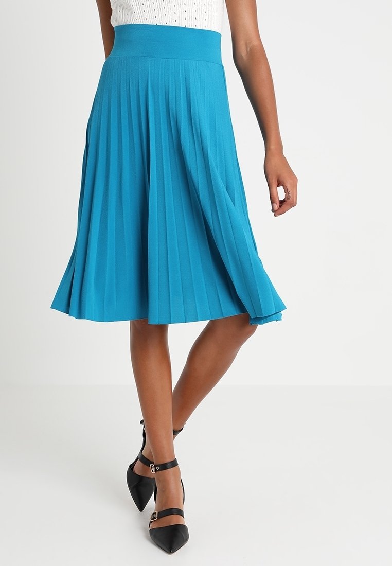 Une jupe plissée d'un turquoise éclatant, en tissu lisse avec une taille haute. La personne la porte avec des talons noirs à bride cheville.