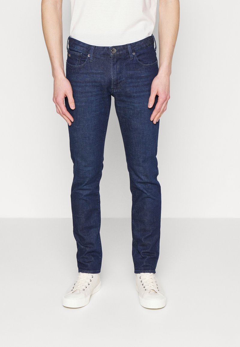 Dark blue denim skinny jeans with a button and zipper closure, five pockets, and subtle stitching details, paired with white sneakers.