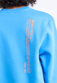 Blå sweatshirt lavet af blødt stof, med orange tekst, der angiver byer samt Nike-logoet langs ryggen. Crew neck design.