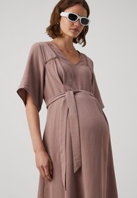 Sukienka ciążowa w miękkim, fakturowanym tkaninie w kolorze mauve, z dekoltem w kształcie litery V, krótkimi rękawami oraz wiązanym detalem w pasie. Udekorowana delikatnym haftem.