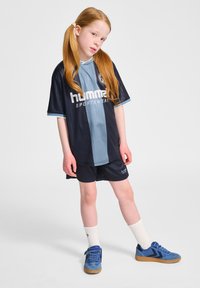 Mörkblå och ljusblå sporttröja med ränder, logotyp och matchande shorts. Modellen har på sig blå sneakers och vita strumpor.