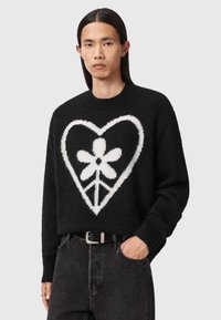 Svart strikkegenser med et stort hvitt hjerte og blomstermønster. Har rund hals og ribbede mansjetter. Brukt med mørke denimjeans.