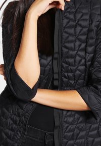 Mujer vestida con una chaqueta acolchada negra de mangas 3/4 dobladas y cerrada hasta la mitad, con un brazo doblado y la mano descansando cerca de la barbilla.