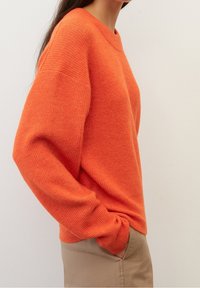 Oranssi neulepaita pyöreällä pääntiellä ja putoavilla harteilla. Resorikuvio, väljä istuvuus, yhdistetty vaalean beigeihin housuihin.