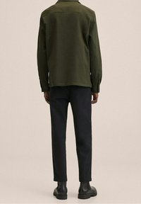 Camicia verde a maniche lunghe con orlo dritto, abbinata a pantaloni scuri su misura e stivali neri, vista da dietro su uno sfondo neutro.