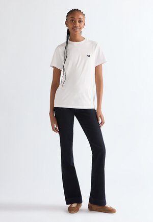 Ung kvinde med fletninger iført en hvid t-shirt med korte ærmer, sorte vide bukser og brune afslappede sko, stående foran en hvid baggrund.