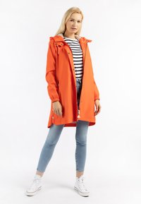 Felgekleurde oranje regenjas met een capuchon, drukknoopsluiting en verstelbare manchetten, gedragen over een zwart-wit gestreept shirt en skinny jeans.