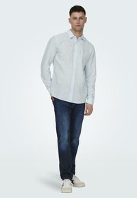 Camicia azzurra chiara con bottoni realizzata in un tessuto testurizzato, abbinata a jeans blu scuro e sneaker bianche. La camicia ha colletto e maniche lunghe.