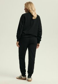 Czarna bluza i pasujące dresy wykonane z miękkiego materiału, z prążkowanymi mankietami i elastycznym paskiem w talii. Na nogach są sneakersy w paski.