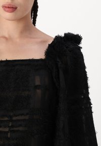 Schwarzes, transparentes Kleid mit strukturiertem Stoff und fransigen Akzenten sowie herzförmigem Ausschnitt. Lange Puffärmel sorgen für Volumen.