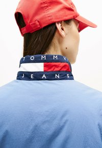 Jasnoniebieska kurtka z granatowym kołnierzem z tekstem "TOMMY JEANS" na białym tle, akcentowana czerwoną i białą paskiem. Czerwona czapka powyżej.