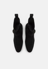 Ett par svarta mockaskor med ankelsnörning, sidobälte och dragkedja, sedd ovanifrån på en vit bakgrund.