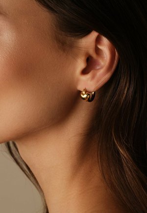 Close-up van het oor van een vrouw die een kleine, glanzende, gouden oorring in de vorm van een hart draagt, met lang donkerbruin haar zichtbaar.