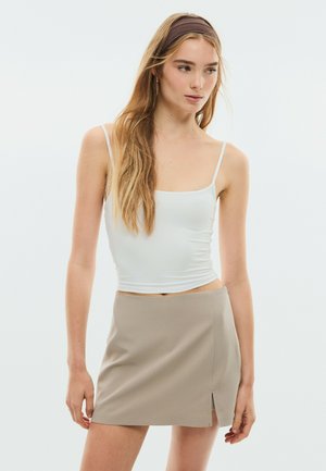 Jeune femme aux longs cheveux blonds portant un top blanc à fines bretelles, une jupe beige avec une fente à l’avant et un bandeau marron, regardant sur le côté.