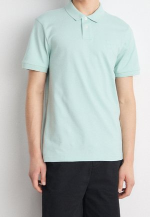 Personne portant un polo vert clair à manches courtes avec deux boutons et un pantalon noir, se tenant devant un fond blanc uni.