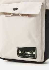 Tasca frontale di una borsa in tessuto beige e nero con bottone a scatto nero e etichetta del marchio Columbia che leggi "Dal 1938" nell'angolo in basso a destra.