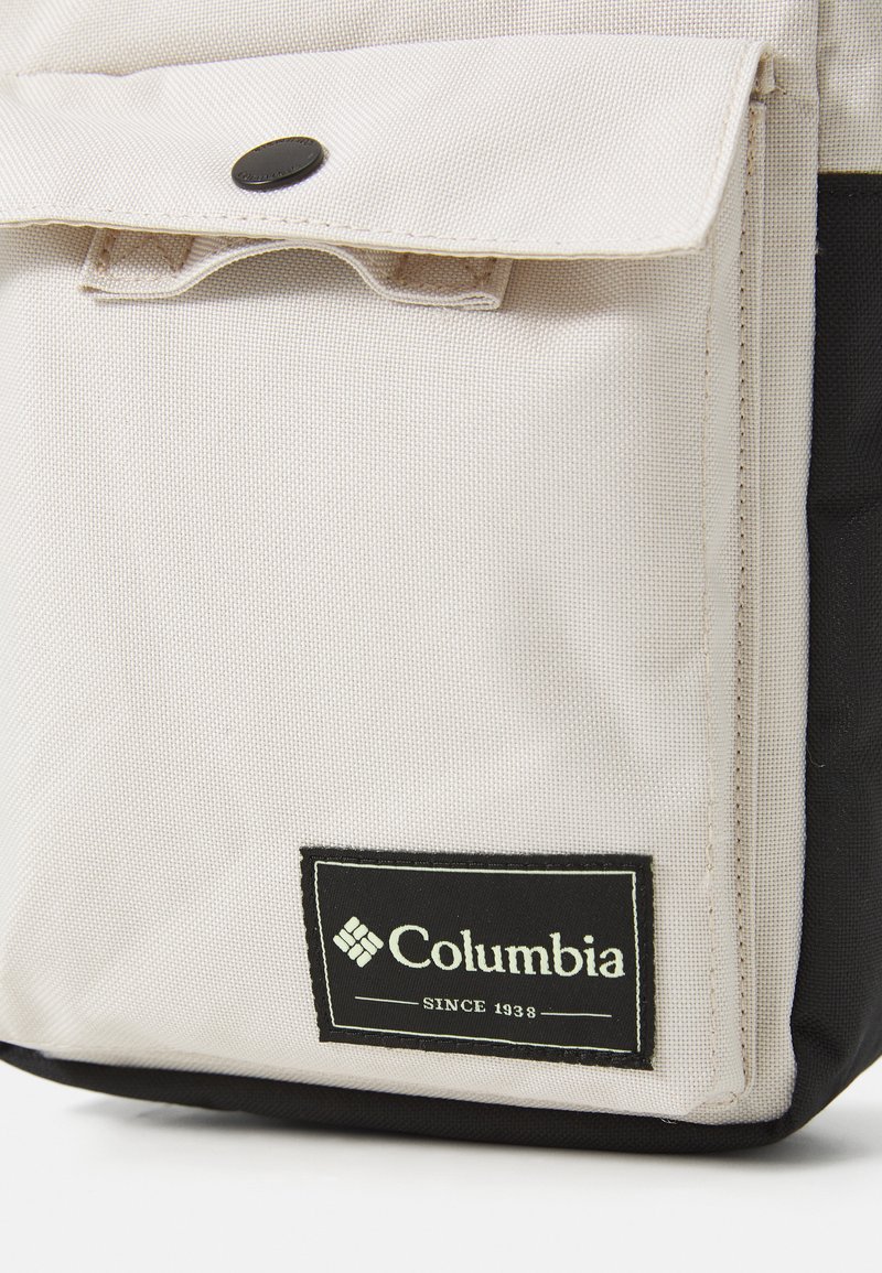 Bolsillo frontal de una bolsa de tela beige y negra con botón a presión negro y etiqueta de la marca Columbia que dice "Desde 1938" en la esquina inferior derecha.