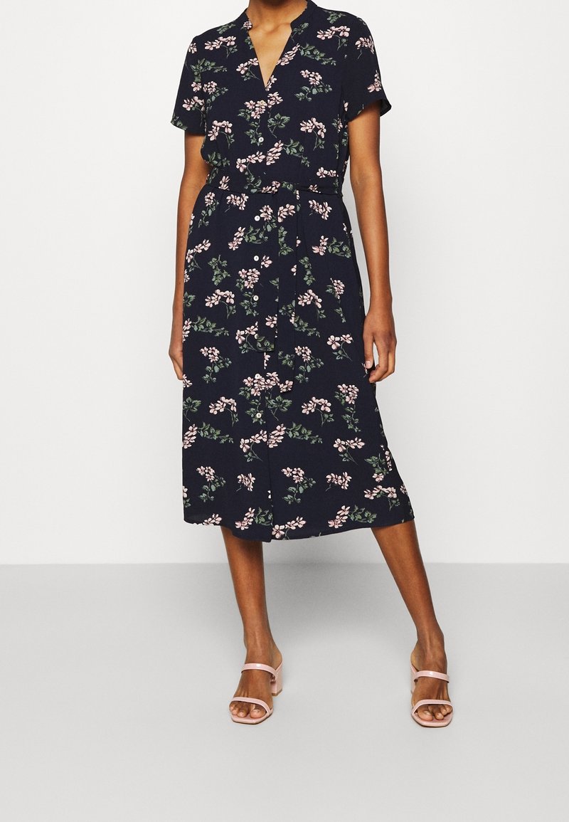 Donna che indossa un abito midi blu navy con stampa floreale rosa, maniche corte, bottone frontale, cintura in vita e sandali con tacco rosa chiaro.