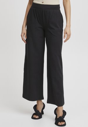 Pantalon large noir en tissu doux, doté d'une taille élastique pour plus de confort. Associé à des talons noirs avec des accents en nœud.