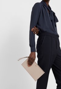 Žena v námořnicky modré blůze a kalhotách drží béžovou kabelku s náramkem na zápěstí na bílém pozadí.