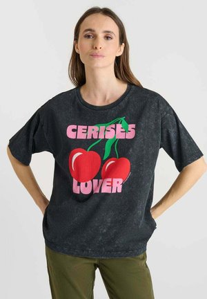 Vrouw met lang bruin haar, gekleed in een zwarte oversized t-shirt met kersen en de tekst "CERISES LOVER", gecombineerd met olijfgroene broeken.