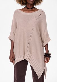 Beige oversized gebreide trui met een geribde onderrand, voorzien van een diagonale lijn van oogjesdetails en opgerolde mouwen. Gepaard met donkerbruine broeken.