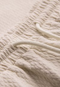 Gros plan d’un tissu beige texturé avec ceinture élastique et cordon, montrant des coutures détaillées et des plis doux du matériau.